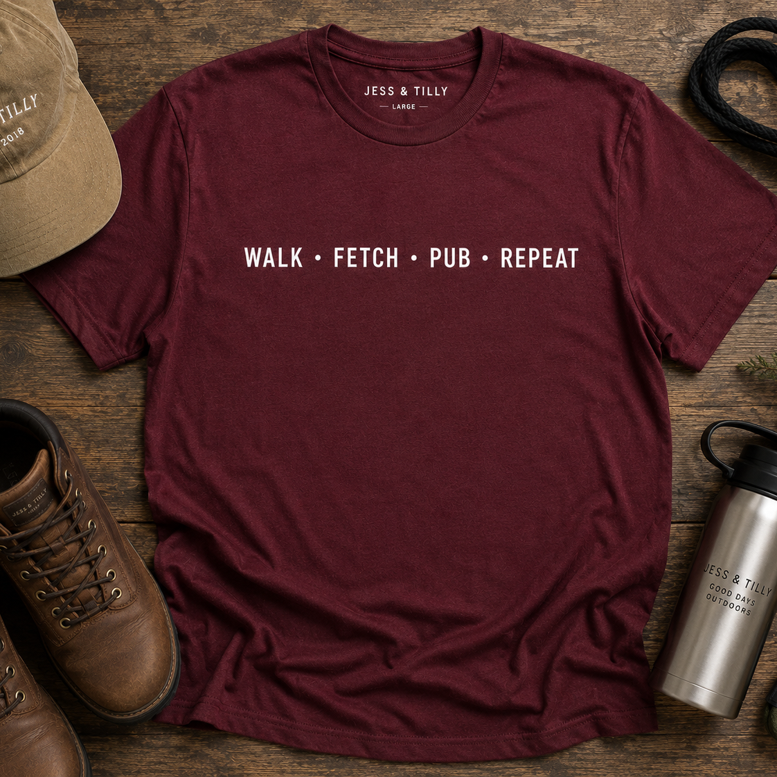 Maroon t-shirt with 'Walk Fetch Pub Repeat' text on a wooden surface with boots, cap, and water bottle.