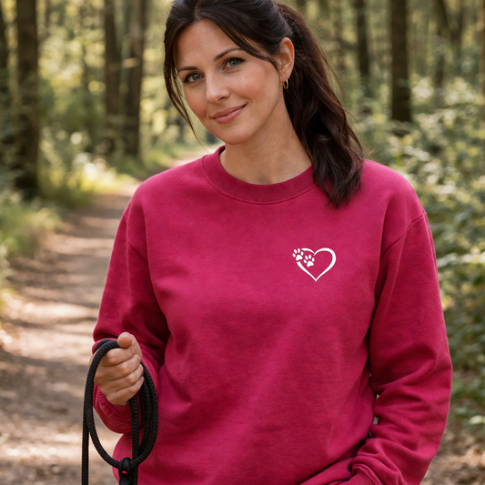 Woman in a pink sweatshirt with a heart and paw print design, holding a dog leash in a forest.