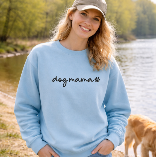 Woman wearing a light blue sweatshirt with 'dogmama' text, standing by a body of water with a dog.