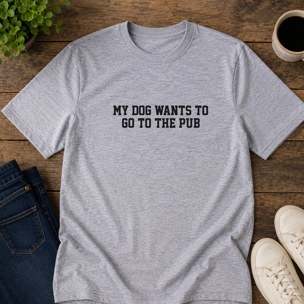 Gray t-shirt with 'My dog wants to go to the pub' text on a wooden surface with shoes and a plant.