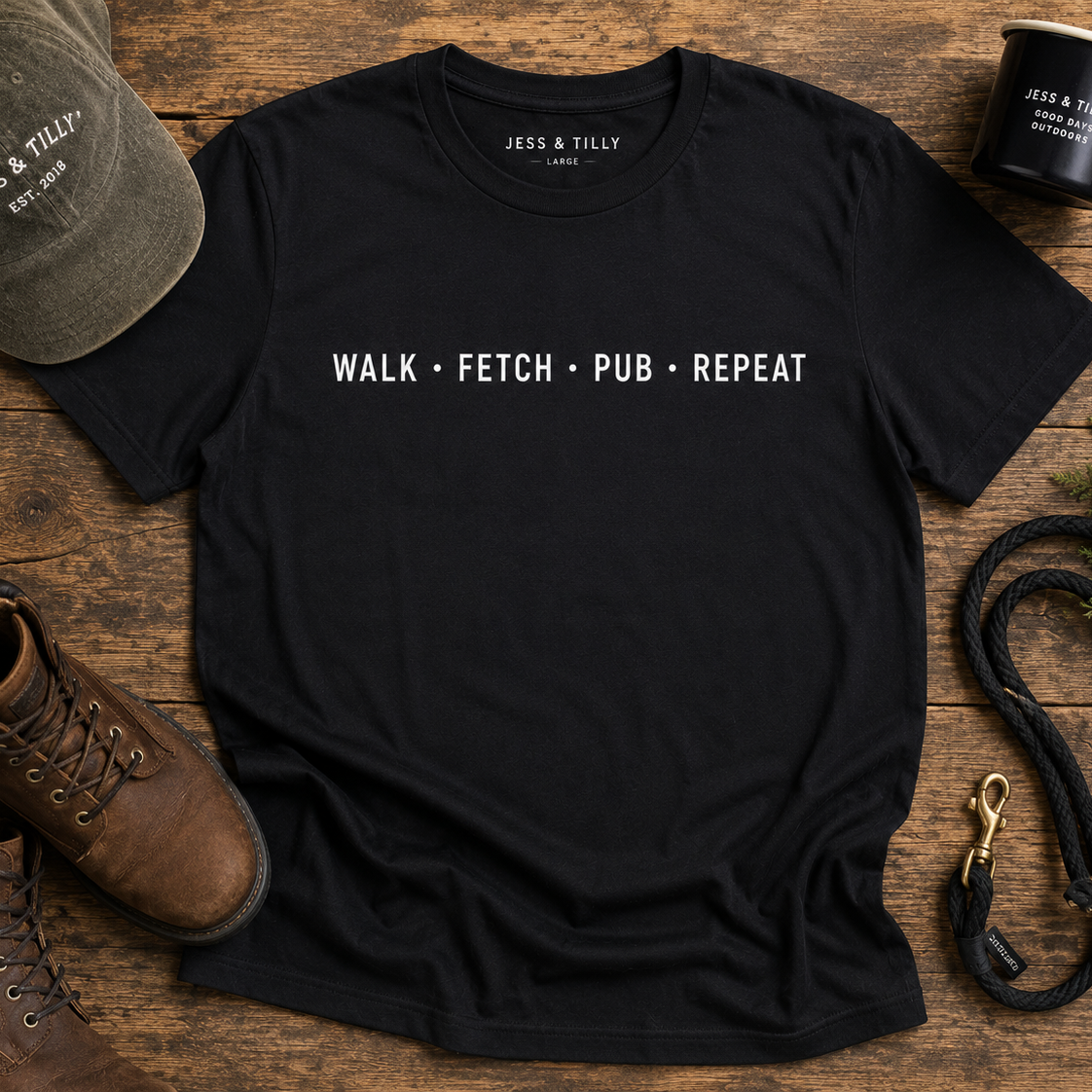 Black t-shirt with 'WALK • FETCH • PUB • REPEAT' text on a wooden surface with a cap, mug, and boots.