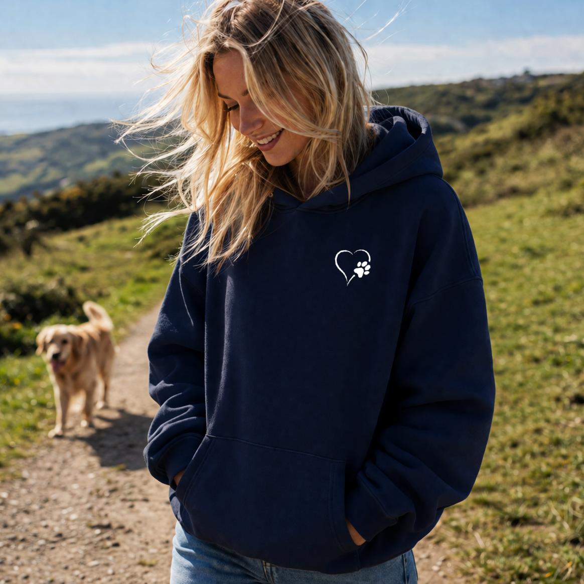 Woman wearing a navy hoodie with a heart and paw print design, standing outdoors with a dog in the background.