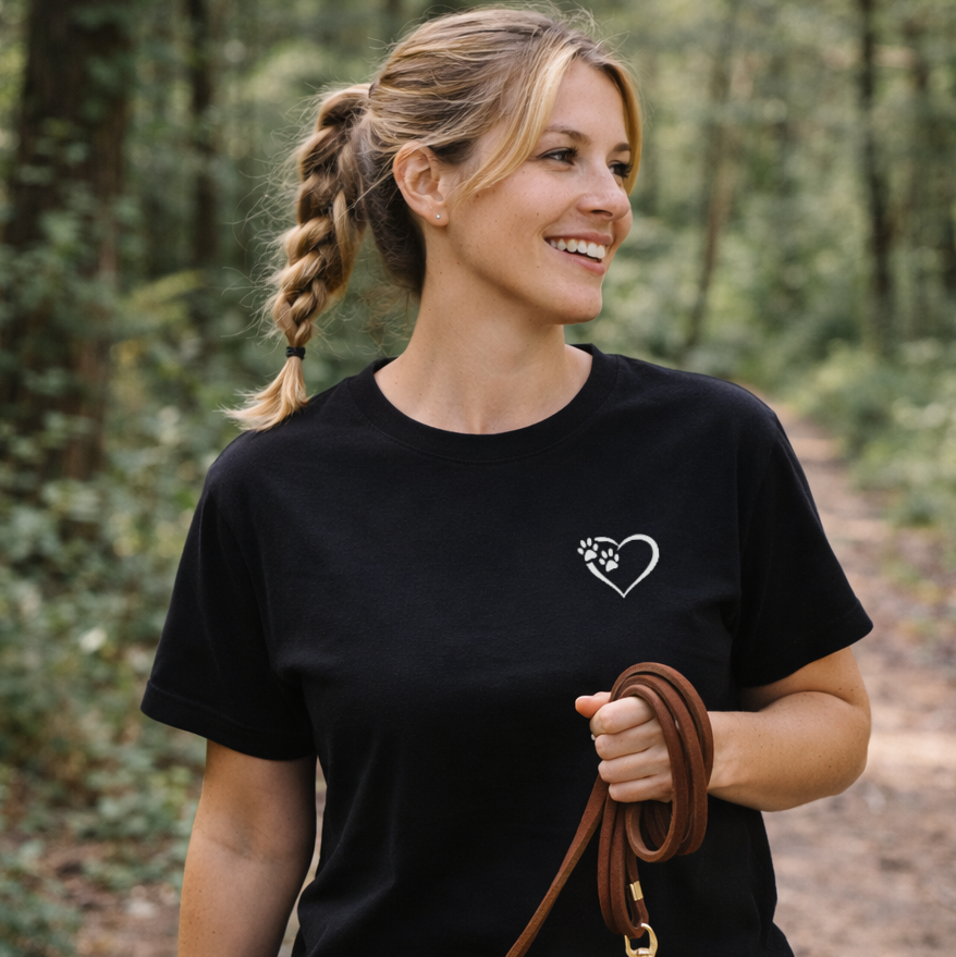 Woman in a black t-shirt with a heart and paw print design, holding a leather leash in a forest setting.