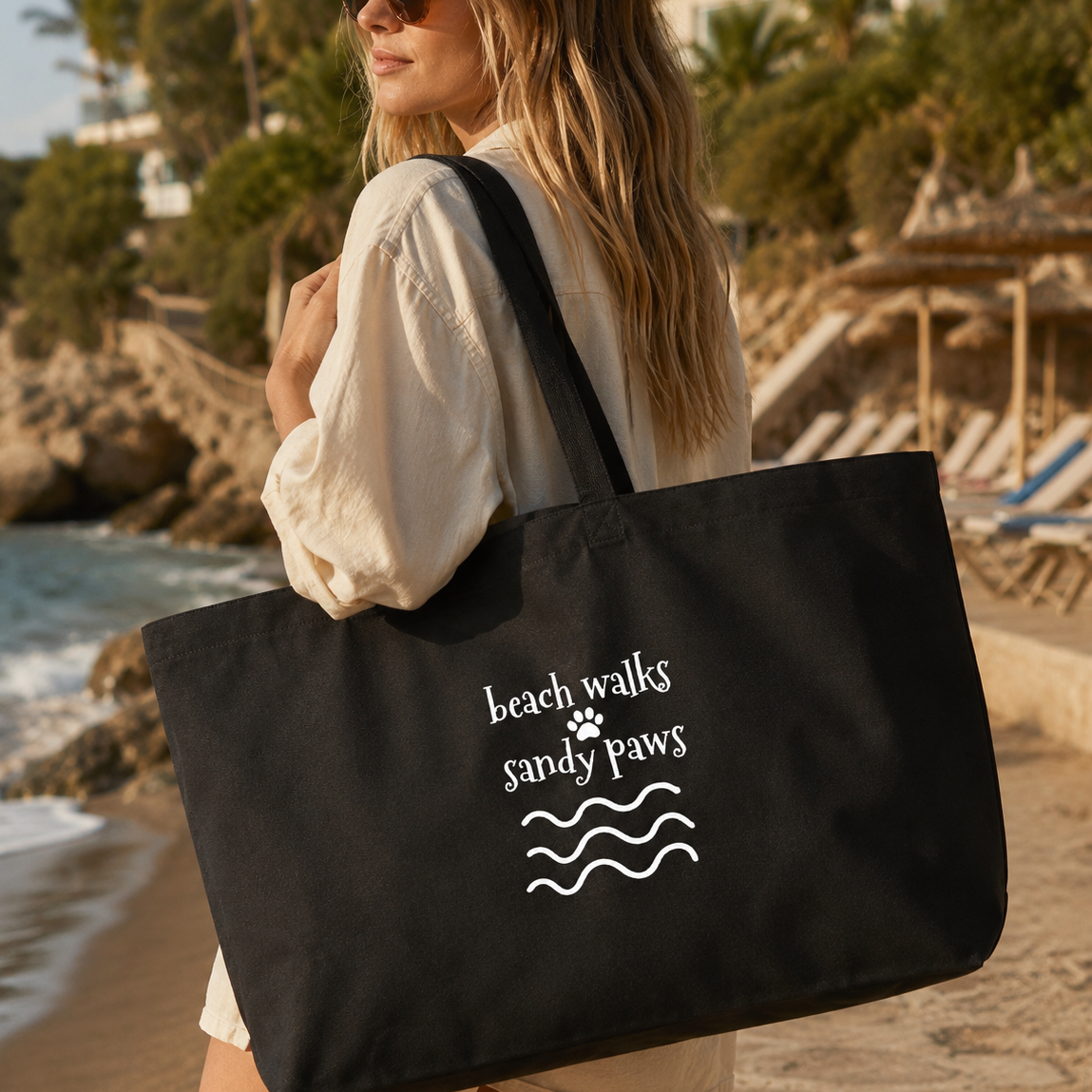 A woman on a beach carries a large black tote bag with white text reading 'beach walks sandy paws' and a paw print above wavy lines. The background shows sand, ocean, palm trees, and beach umbrellas.