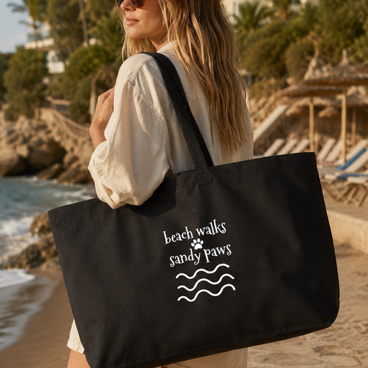 A woman on a beach carries a large black tote bag with white text reading 'beach walks sandy paws' and a paw print above wavy lines. The background shows sand, ocean, palm trees, and beach umbrellas.