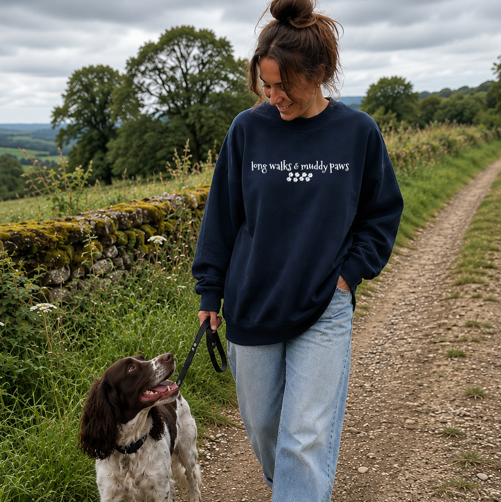 A woman walking a brown and white dog on a dirt path outdoors, wearing a navy blue sweatshirt with the white text 'pet walks & muddy paws' and a small graphic, paired with light blue jeans.
