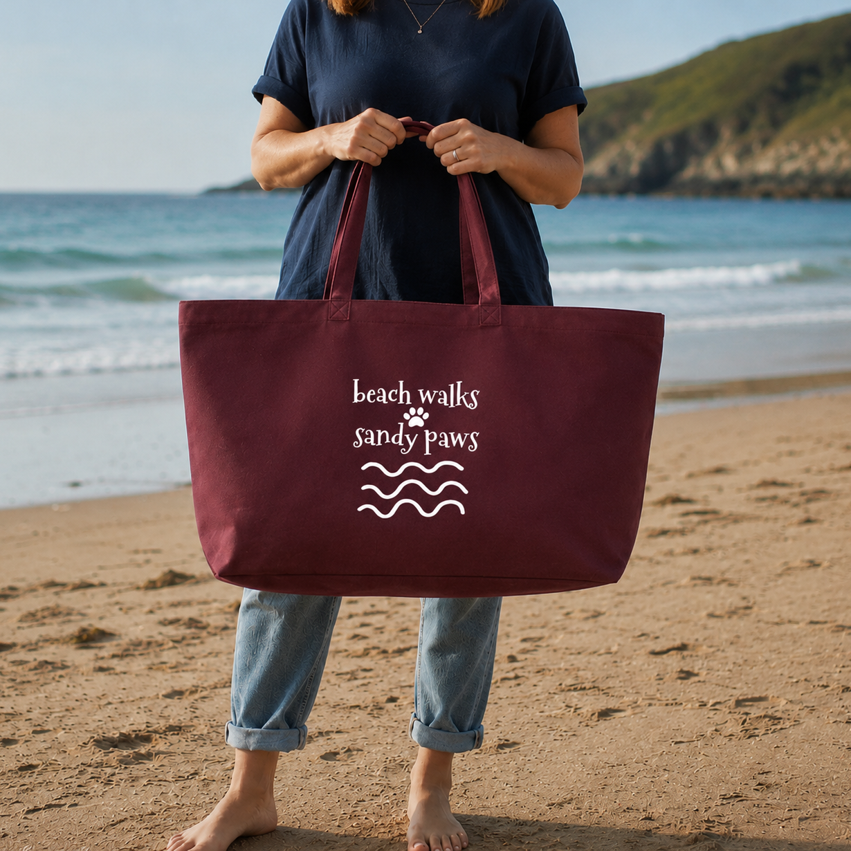 A person standing on a sandy beach holds a large maroon tote bag with white text reading 'beach walks sandy paws' and a small paw print motif. The ocean and shoreline are visible in the background.