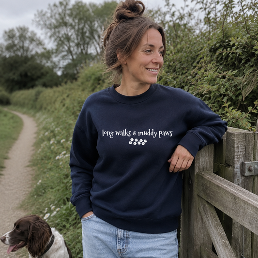 A woman wearing a navy blue crewneck sweatshirt with the white text 'long walks & muddy paws' and paw prints, leaning on a wooden fence outdoors next to a dog.